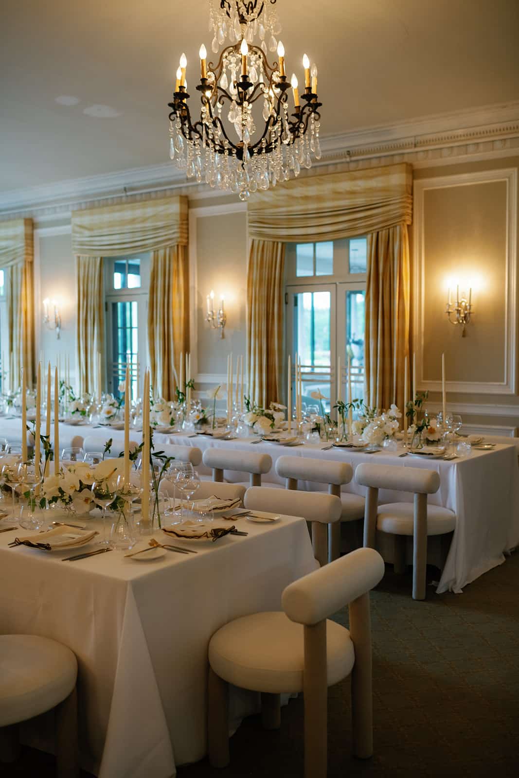 Dinner party tables set in a creamy ballroom with flowers, linens, taper candles, napkins, and chairs in monochrome neutrals.